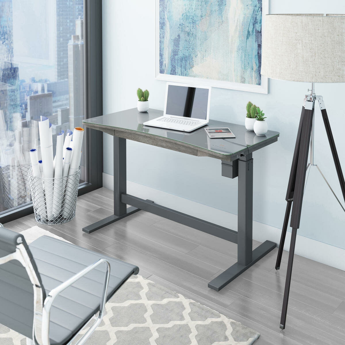 People benefit from electronic standing desks when they need to work from home. A brown standing desk is placed next to a white wall with serene art and a white lamp.